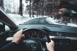 mans hands on the black steering wheel of a black car driving over a cleared road in snowy conditions.