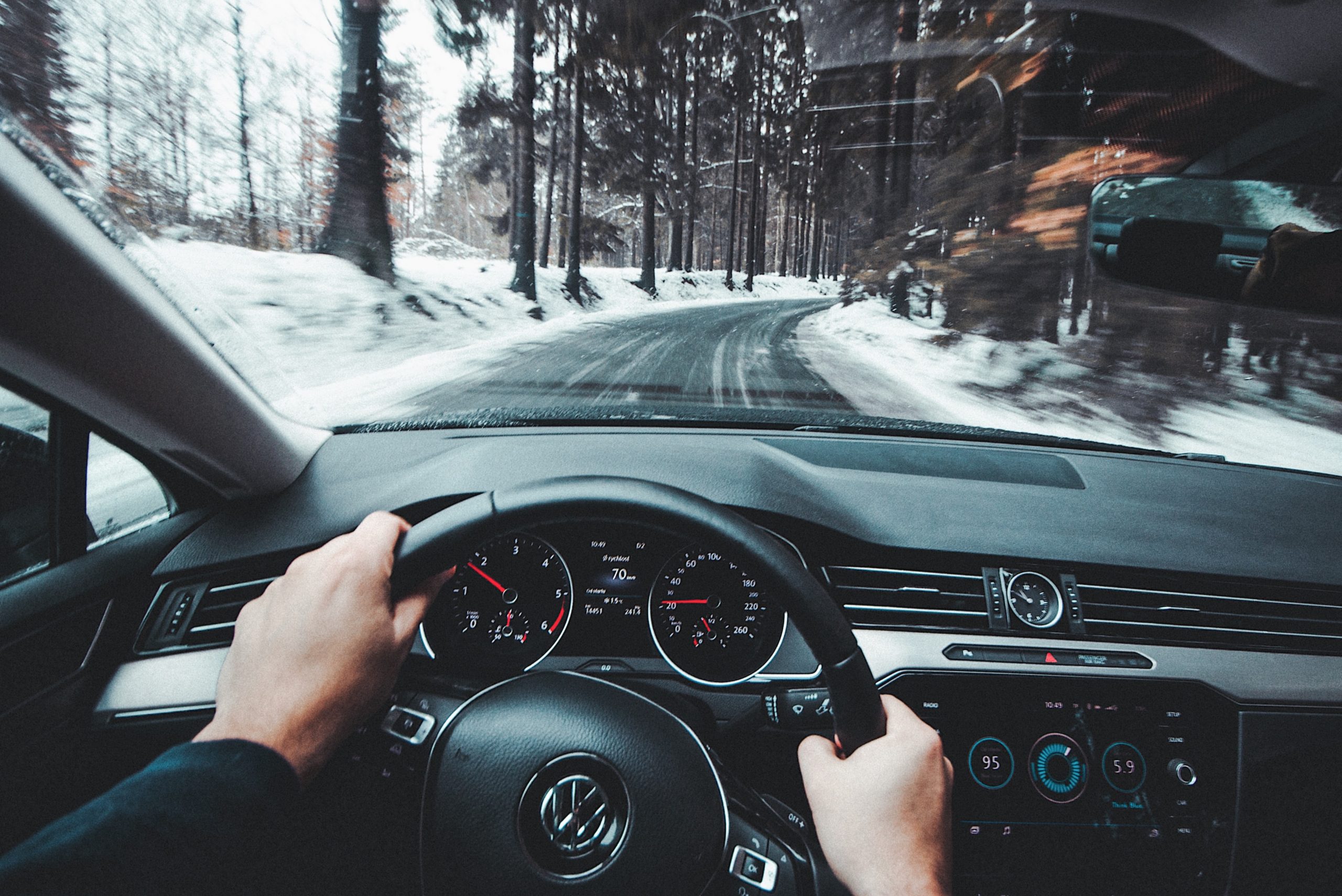 mans hands on the black steering wheel of a black car driving over a cleared road in snowy conditions.