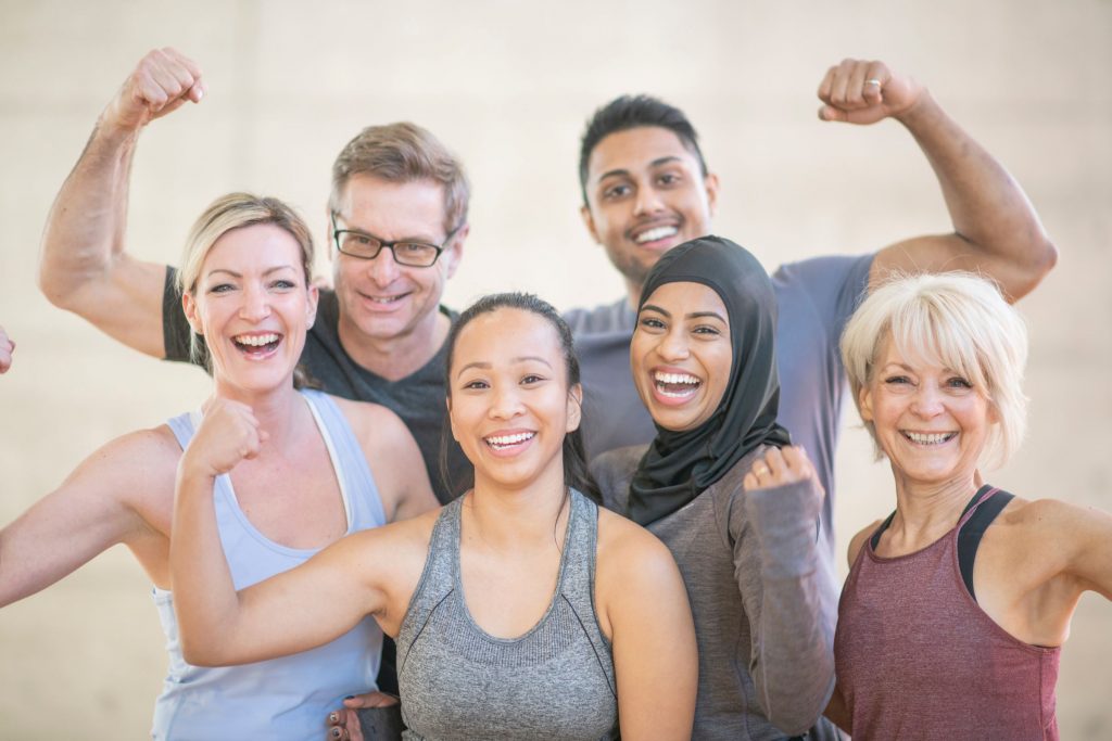 Group of 5 people, men and women, in pastel and grey colors, holding up one harm in a 'strong' bicep position post-workout
