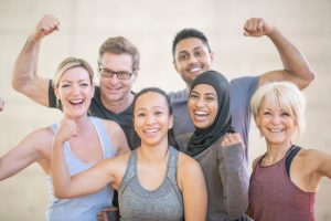 Group of 5 people, men and women, in pastel and grey colors, holding up one harm in a 'strong' bicep position post-workout