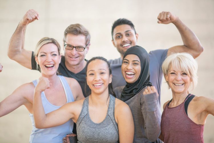 Group of 5 people, men and women, in pastel and grey colors, holding up one harm in a 'strong' bicep position post-workout